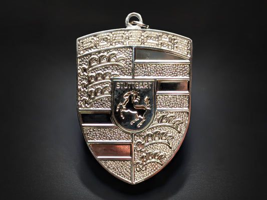 Porsche silver badge keychain featuring a monochrome 3D metallic crest with a textured background and Stuttgart horse detail.