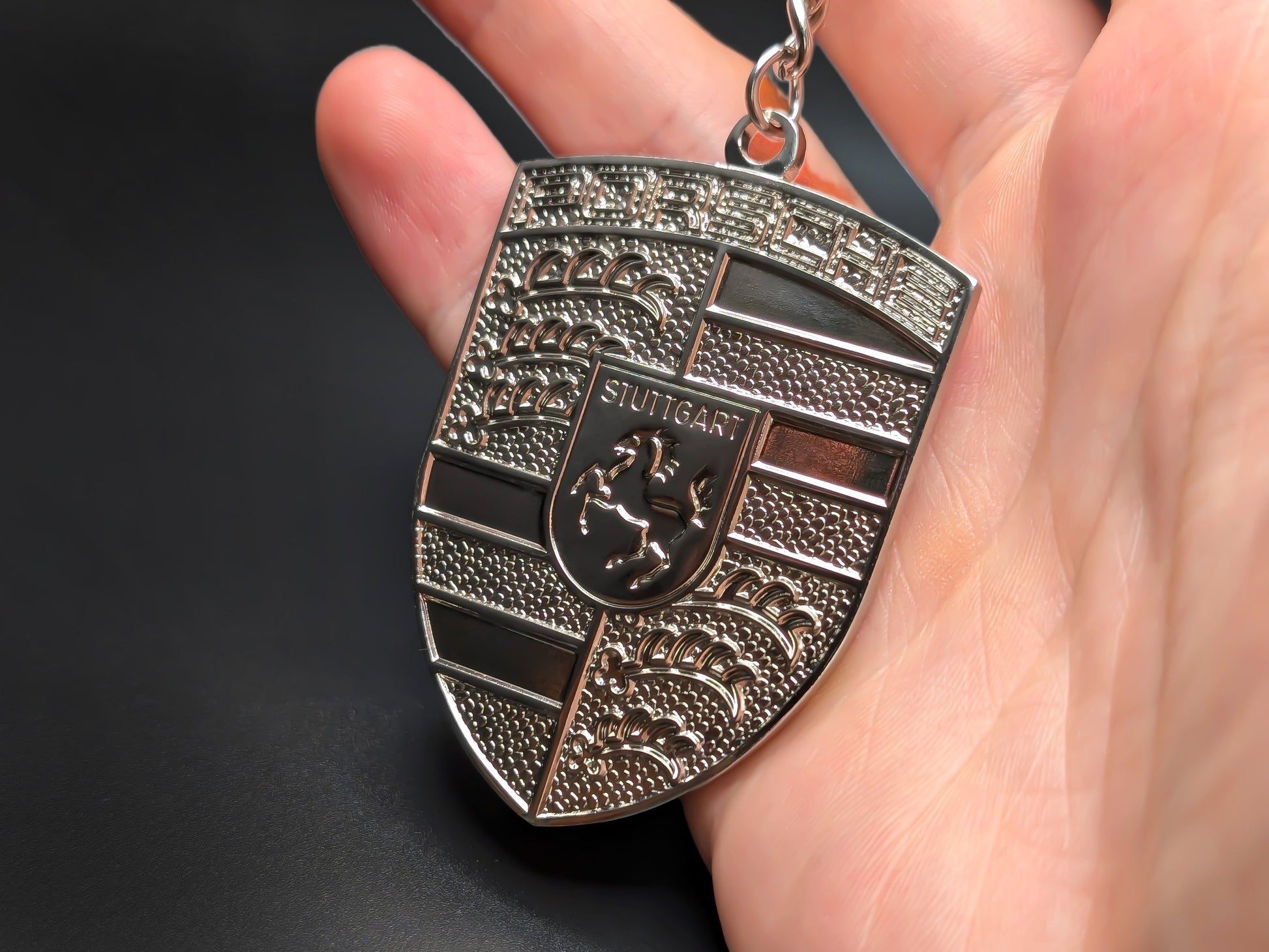 Hand holding a monochrome Porsche silver badge keychain to demonstrate the premium metallic weight and subtle 3D textures.