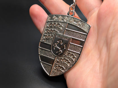 Hand holding a monochrome Porsche silver badge keychain to demonstrate the premium metallic weight and subtle 3D textures.