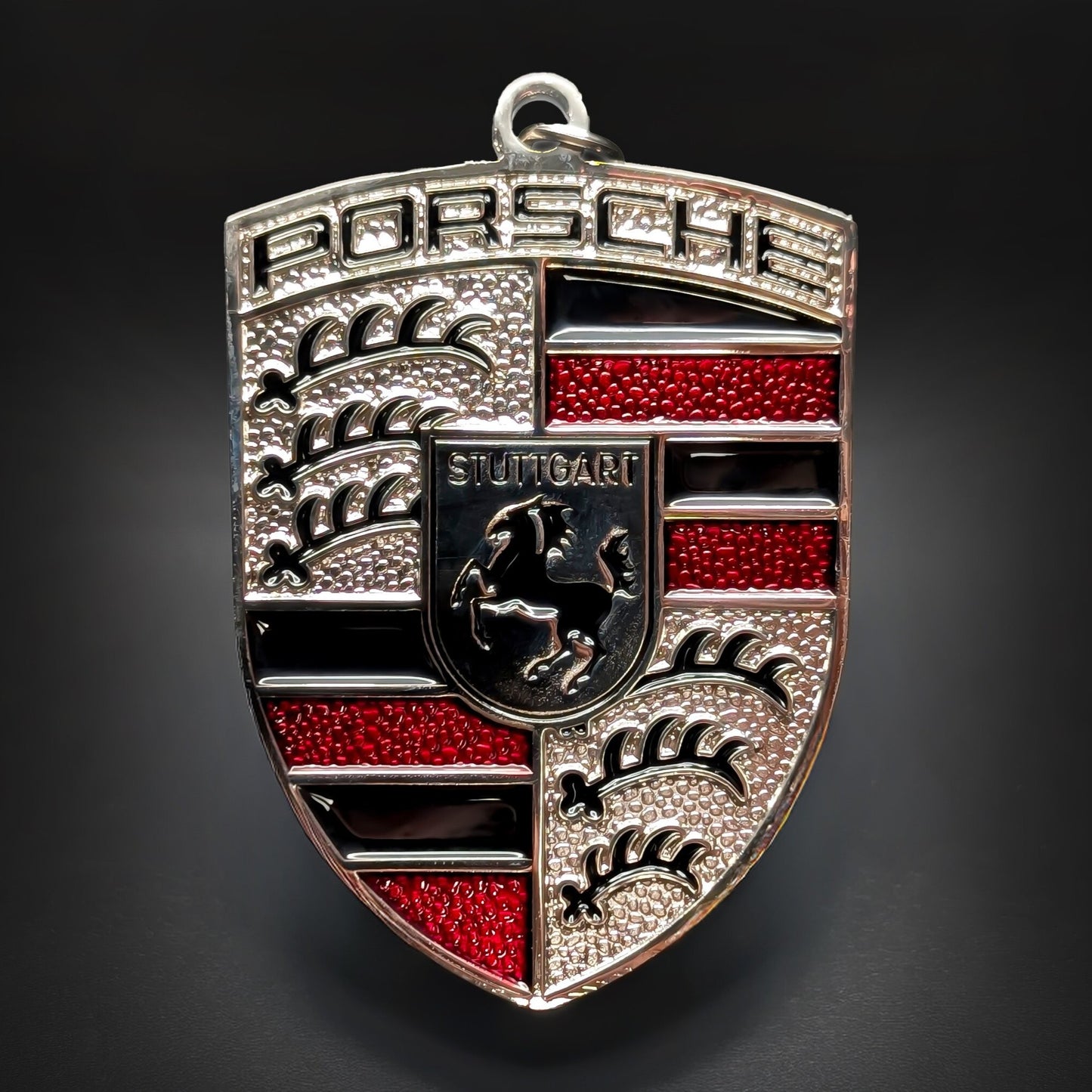 Double-sided metal Porsche crest badge keychain in silver with red enamel accents and black bands, a central STUTTGART shield with a black horse, and textured quarters, on a dark surface.