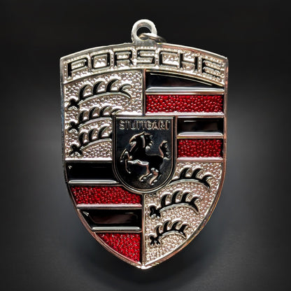 Double-sided metal Porsche crest badge keychain in silver with red enamel accents and black bands, a central STUTTGART shield with a black horse, and textured quarters, on a dark surface.