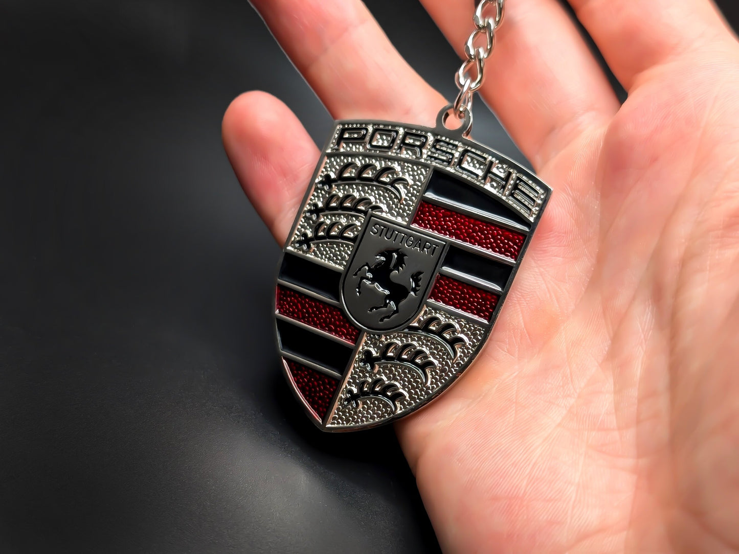 Hand holding a 3D Porsche red badge keychain to demonstrate the premium metallic weight and textured enamel details.