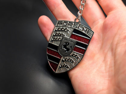 Hand holding a 3D Porsche red badge keychain to demonstrate the premium metallic weight and textured enamel details.