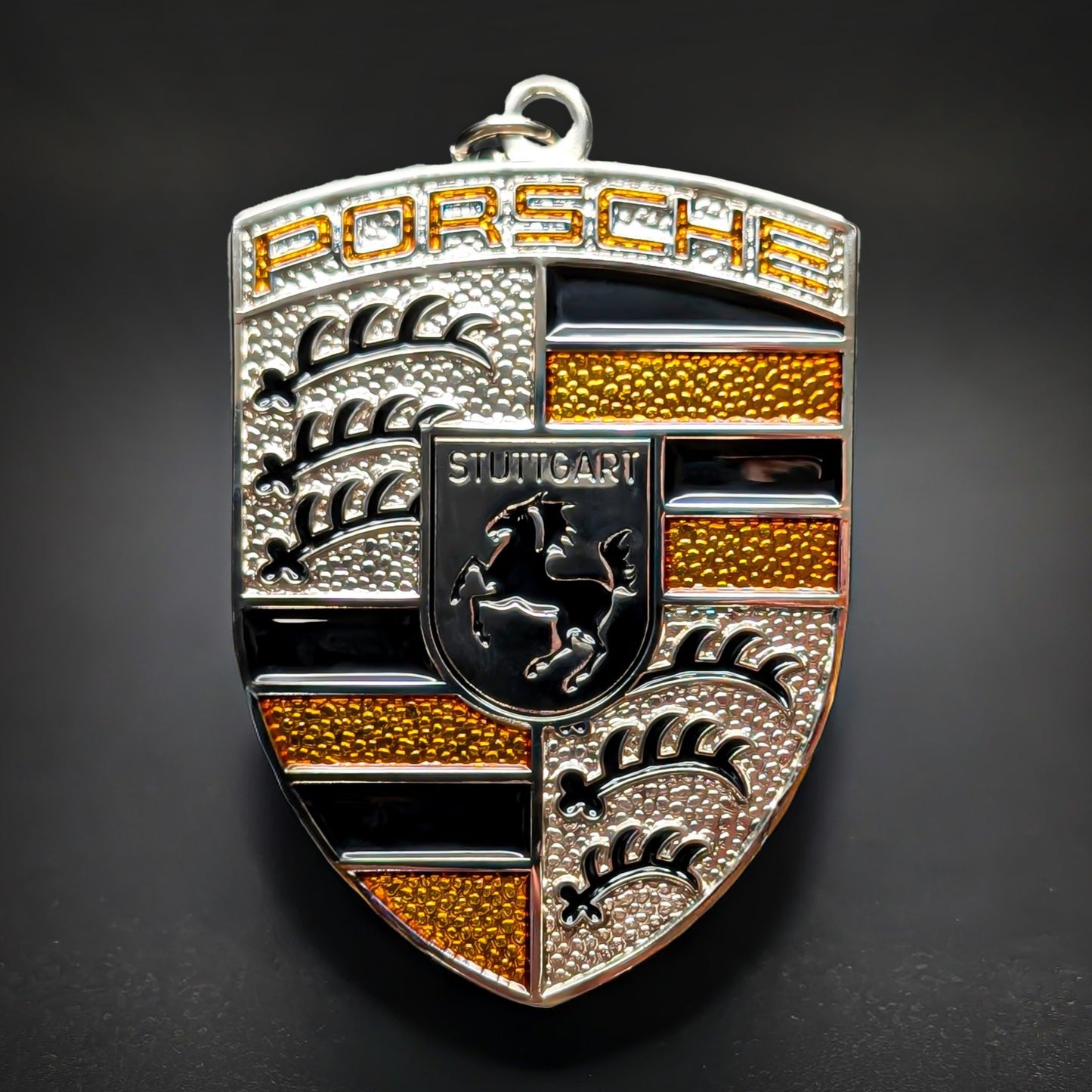 Double-sided metal Porsche crest badge keychain in silver with amber-yellow enamel accents, an amber-tinted PORSCHE wordmark, and a central STUTTGART shield with a black horse, on a dark surface.