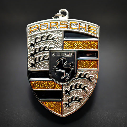 Double-sided metal Porsche crest badge keychain in silver with amber-yellow enamel accents, an amber-tinted PORSCHE wordmark, and a central STUTTGART shield with a black horse, on a dark surface.