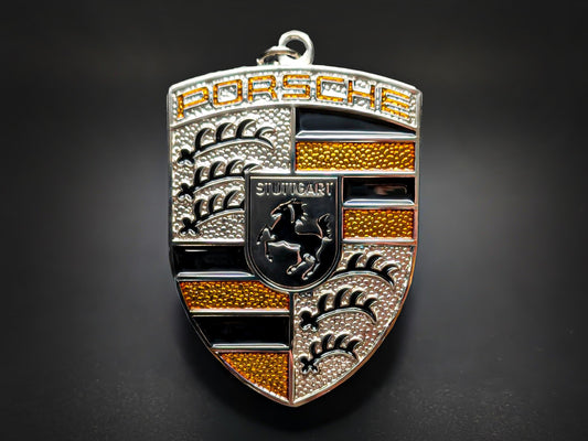 Porsche badge keychain featuring a textured yellow and silver crest with the Stuttgart horse and 3D embossed lettering.