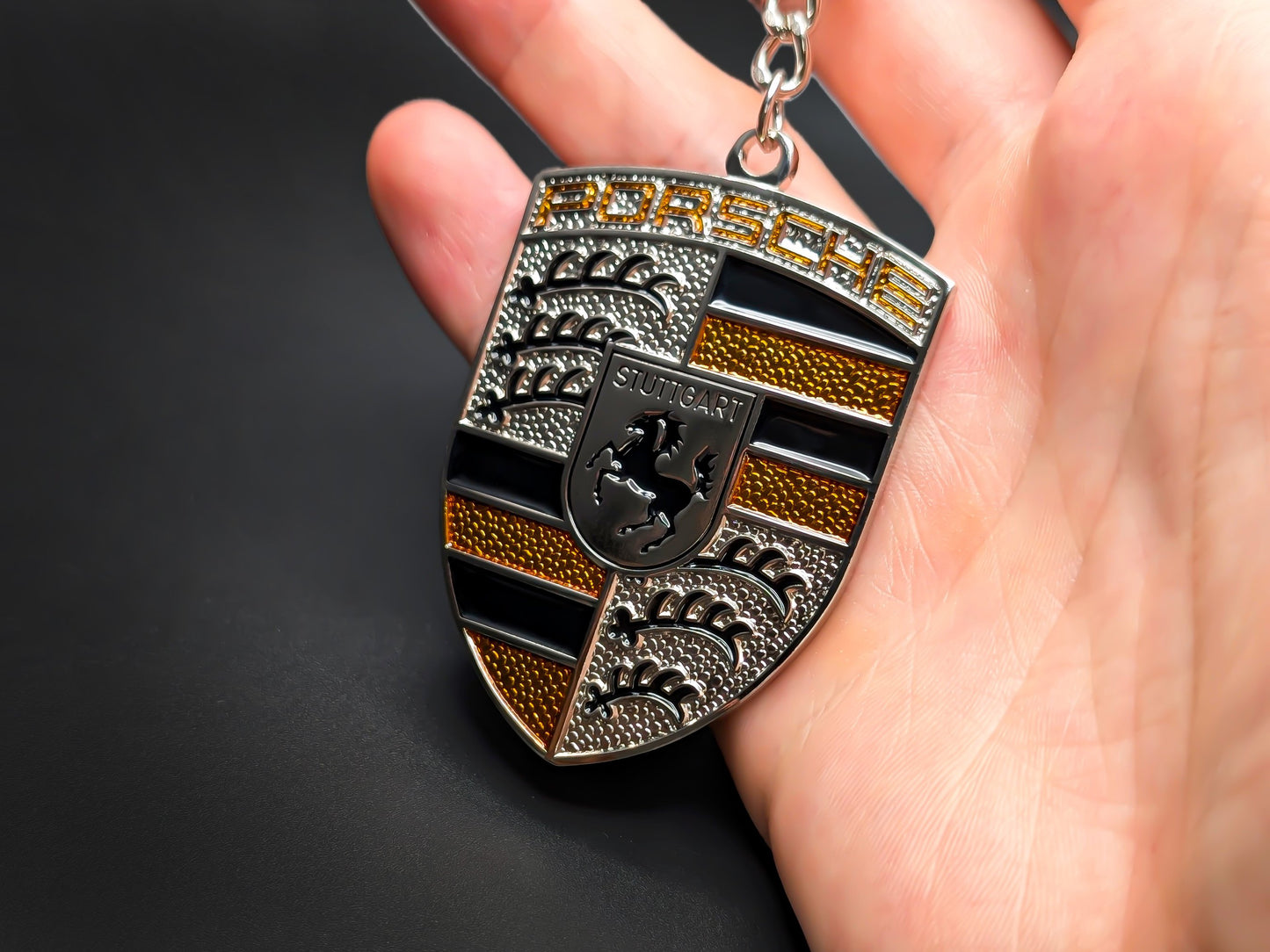 Hand holding a 3D Porsche yellow badge keychain to demonstrate the premium metallic weight and textured amber details.