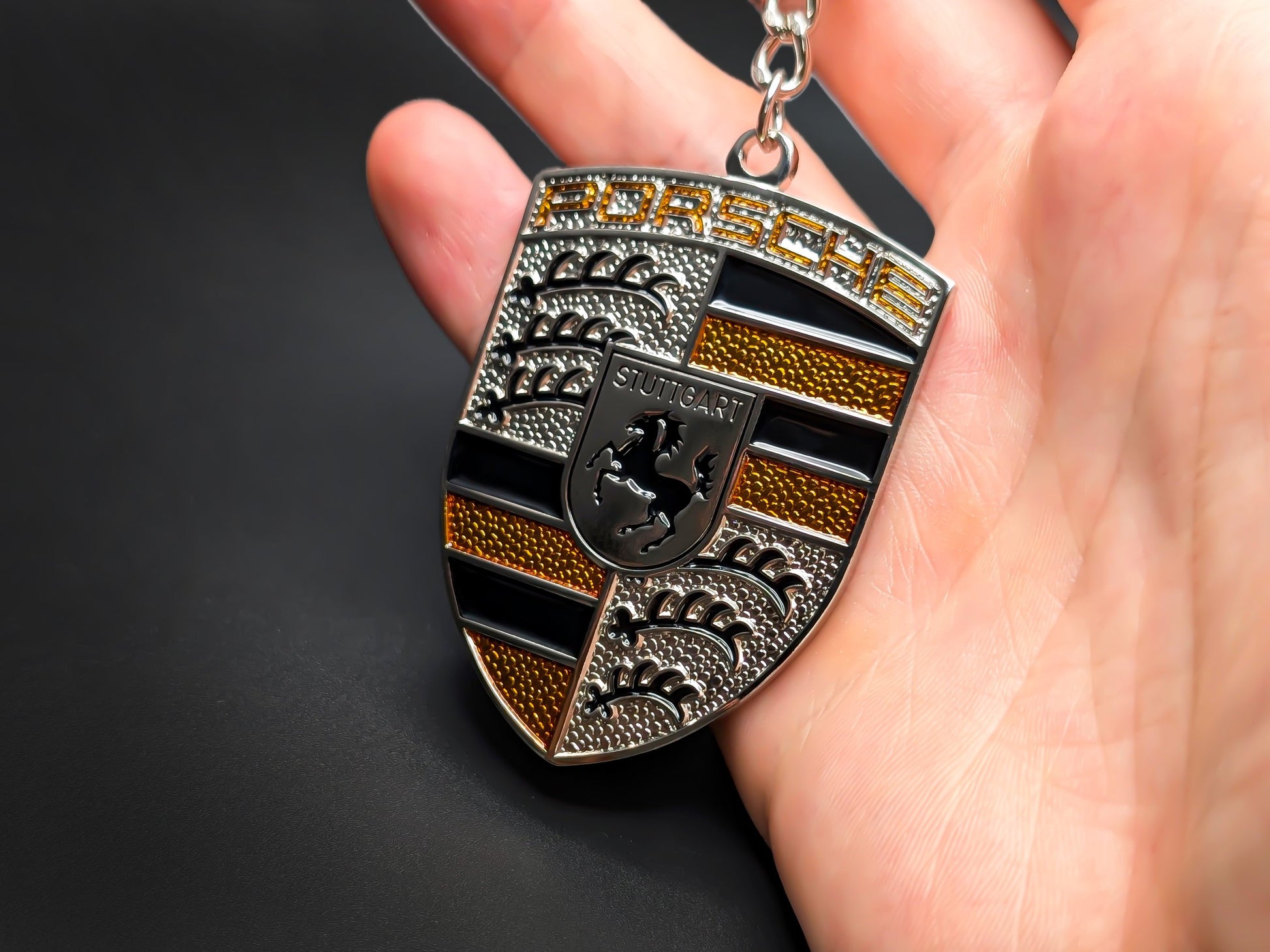 Hand holding a 3D Porsche yellow badge keychain to demonstrate the premium metallic weight and textured amber details.