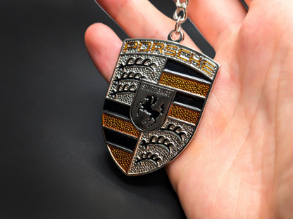 Hand holding a 3D Porsche yellow badge keychain to demonstrate the premium metallic weight and textured amber details.