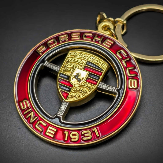 Double-sided metal Porsche badge keychain with a red enamel ring reading PORSCHE CLUB and SINCE 1931 in gold, a gold crest with black horse emblem, gold chain and split ring, on a dark surface.