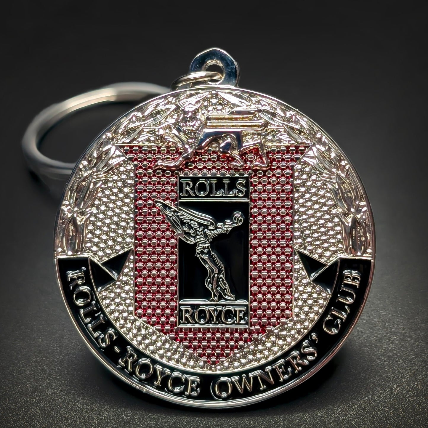 Double-sided metal Rolls-Royce badge keychain with the Spirit of Ecstasy figure on a black and red textured panel, ROLLS-ROYCE OWNERS’ CLUB text at the bottom, silver split ring, on a dark surface.