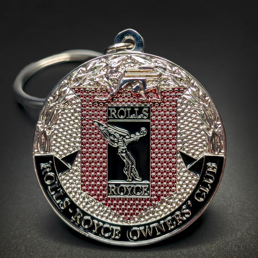 Double-sided metal Rolls-Royce badge keychain with the Spirit of Ecstasy figure on a black and red textured panel, ROLLS-ROYCE OWNERS’ CLUB text at the bottom, silver split ring, on a dark surface.
