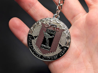 A hand holding the Rolls-Royce Owners' Club badge keychain, highlighting the intricate textures and polished silver-tone finish.