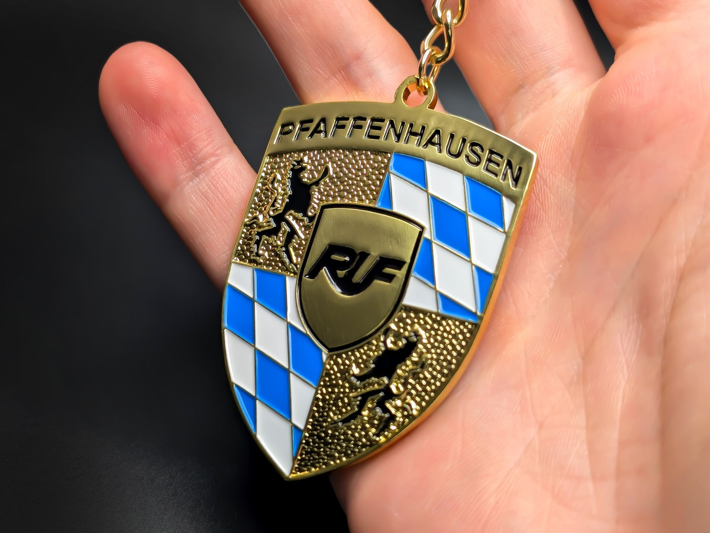 Hand holding a gold RUF Pfaffenhausen enamel badge keychain to demonstrate the premium weight and 3D raised details.