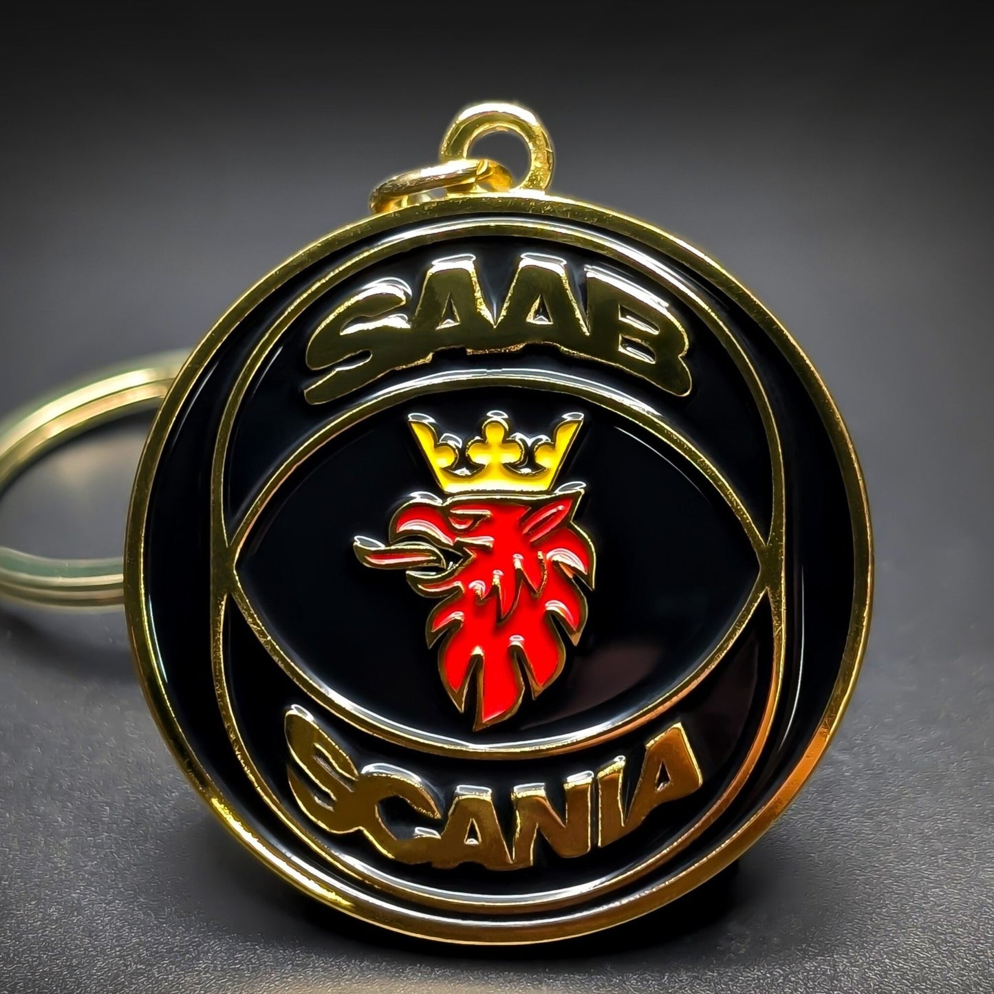 Double-sided metal Saab badge keychain with gold ‘SAAB’ and ‘SCANIA’ lettering framing a red griffin head crest on a glossy black enamel background, gold-tone split ring attached, on a dark surface.