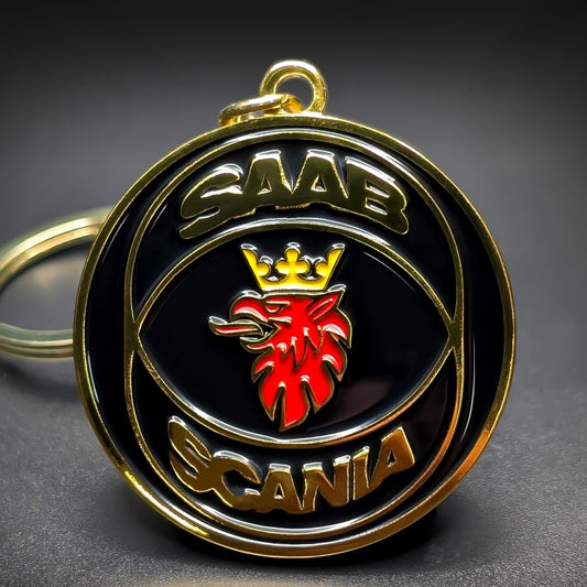 Double-sided metal Saab badge keychain with gold ‘SAAB’ and ‘SCANIA’ lettering framing a red griffin head crest on a glossy black enamel background, gold-tone split ring attached, on a dark surface.