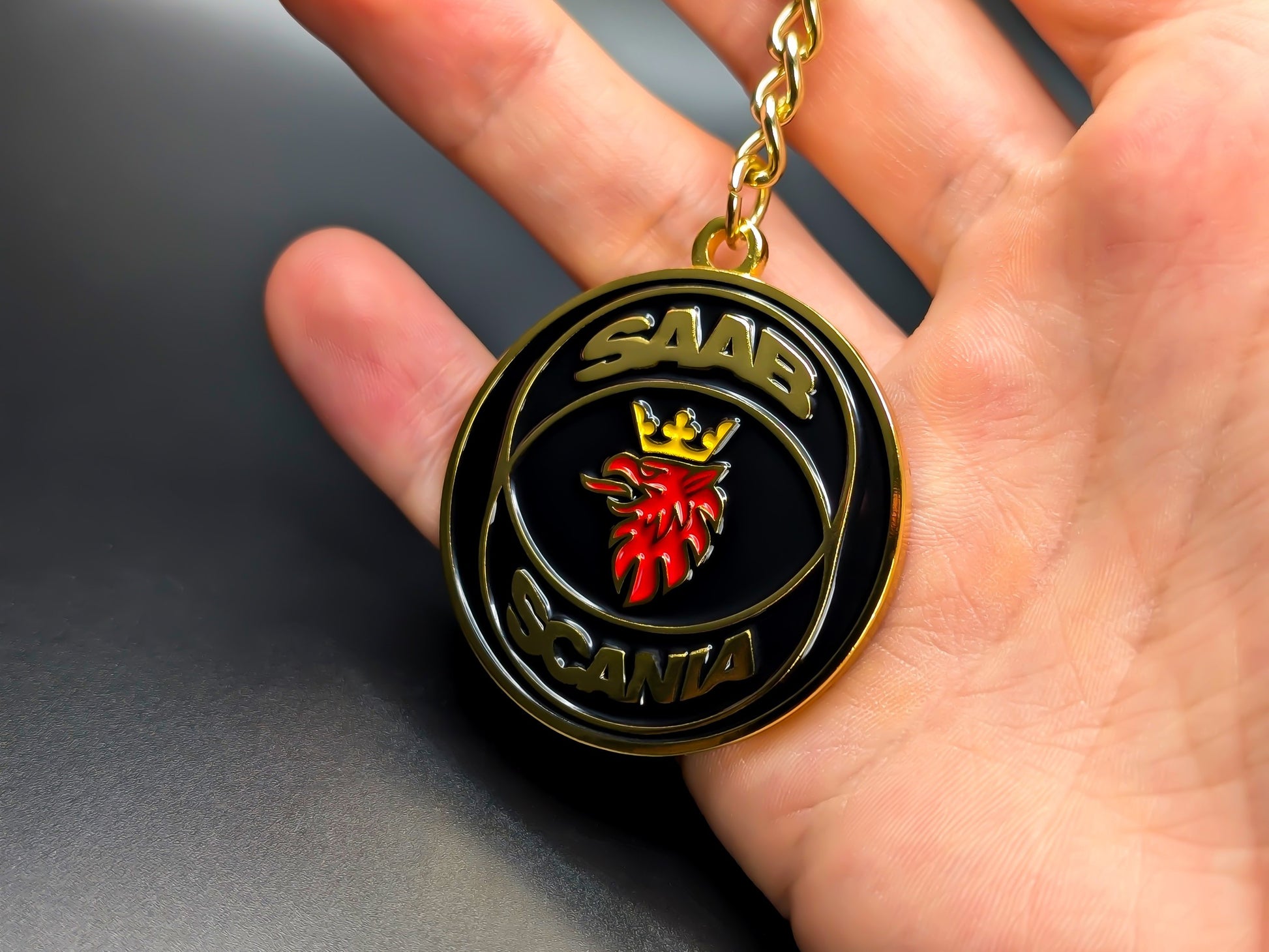 Hand holding a circular Saab Scania black and gold enamel badge keychain to demonstrate its premium metallic weight and detailed finish.