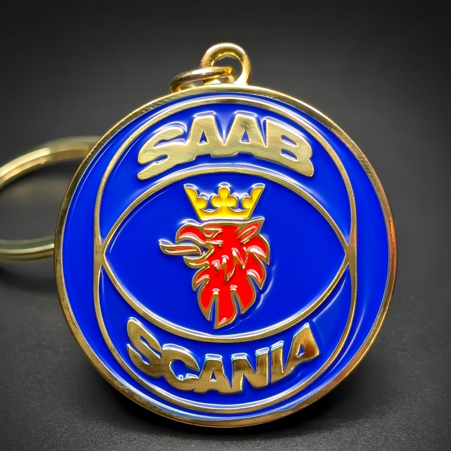 Double-sided metal Saab badge keychain with gold ‘SAAB’ and ‘SCANIA’ lettering framing a red griffin head crest on a blue enamel background, gold-tone split ring attached, on a dark surface.