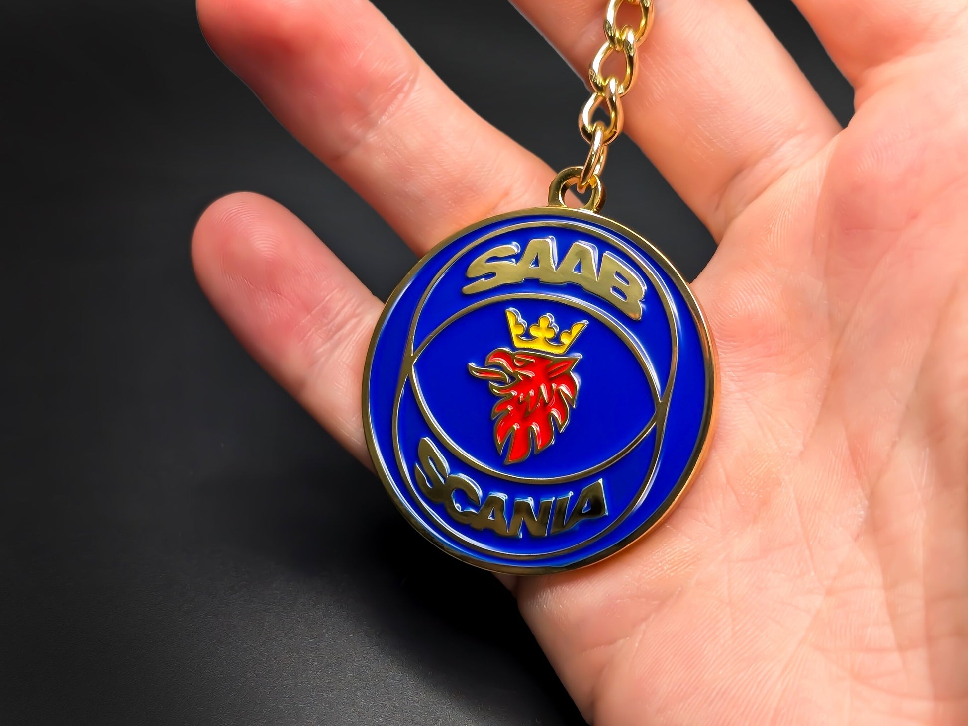 Hand holding a circular Saab Scania original badge keychain to demonstrate the premium metallic weight and vibrant color finish.