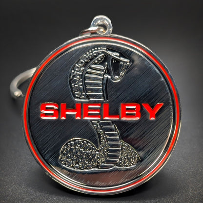 Double-sided metal Shelby badge keychain with a chrome cobra coiled around red SHELBY lettering on a brushed dark gunmetal background, thin red ring border, chrome hook clasp, on a dark surface.