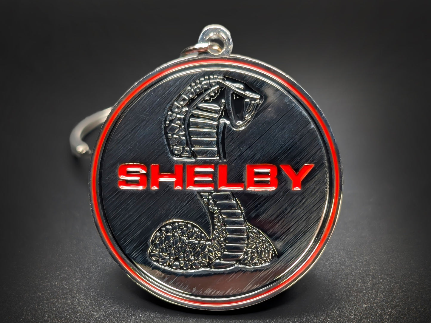 Close-up of a circular metal badge keychain featuring the iconic Shelby Cobra emblem against a high-contrast background.