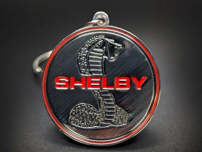 Close-up of a circular metal badge keychain featuring the iconic Shelby Cobra emblem against a high-contrast background.