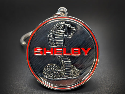 Close-up of a circular metal badge keychain featuring the iconic Shelby Cobra emblem against a high-contrast background.