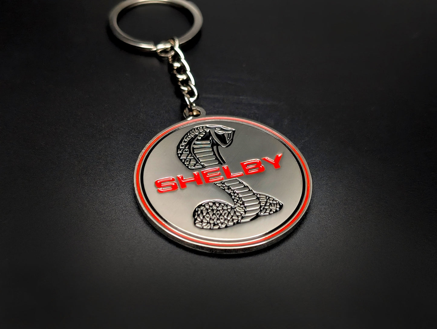A Shelby Cobra badge keychain with a silver chain and ring, showcased on a clean, dark surface.
