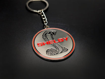 A Shelby Cobra badge keychain with a silver chain and ring, showcased on a clean, dark surface.