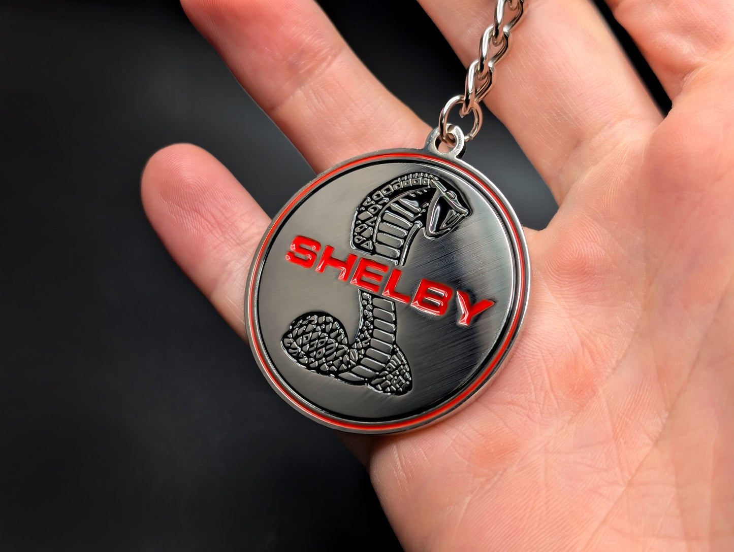 A hand holding the Shelby Cobra badge keychain, highlighting the solid metal construction and detailed emblem design.