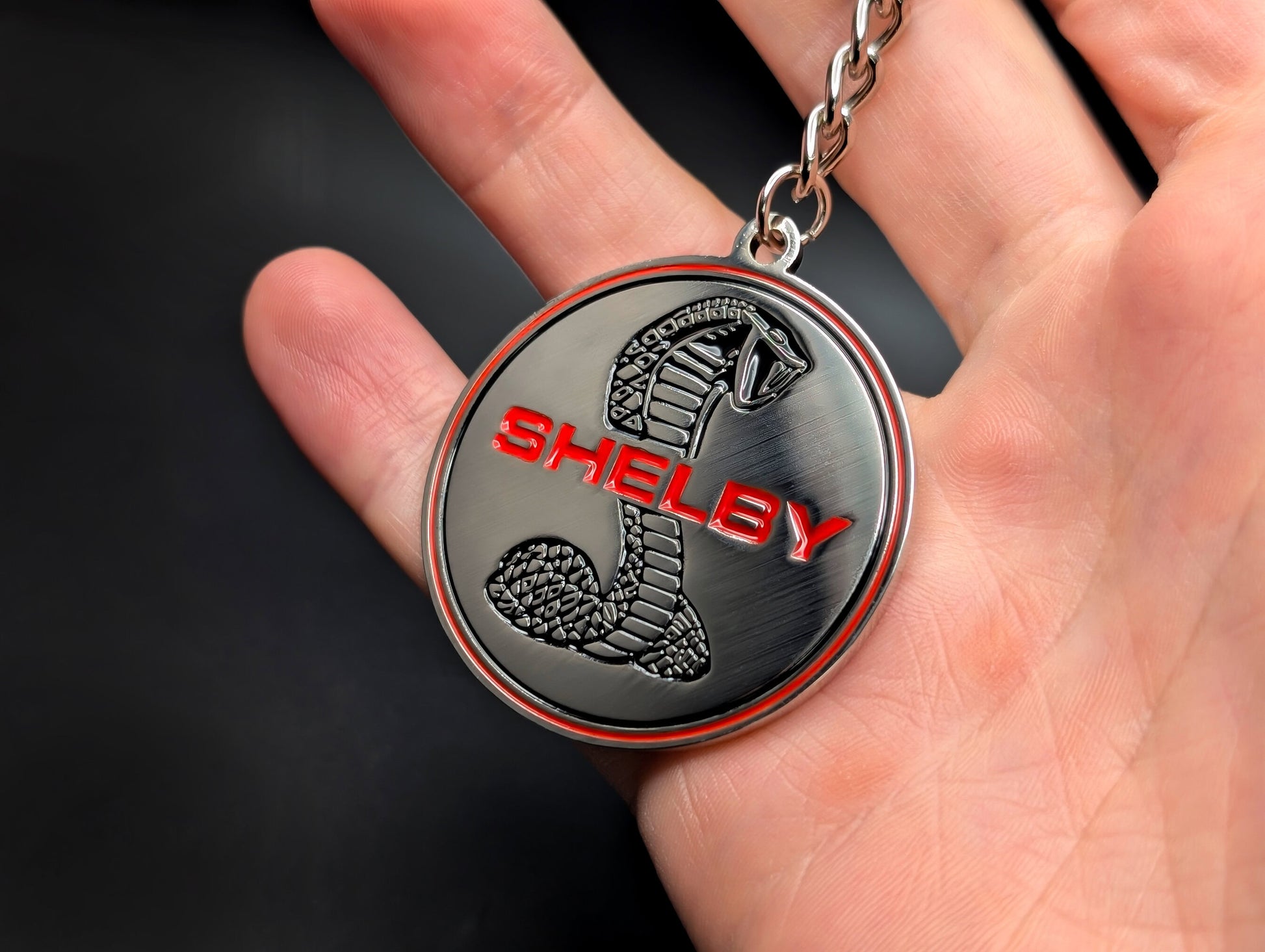 A hand holding the Shelby Cobra badge keychain, highlighting the solid metal construction and detailed emblem design.