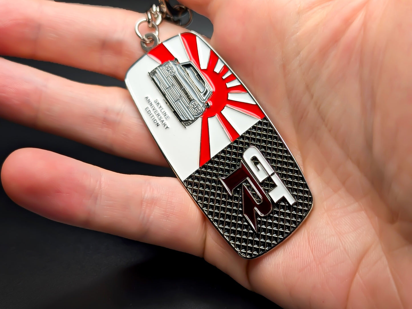 Hand holding the Skyline GT-R anniversary badge keychain to display the polished metal finish and detailed enamel work.