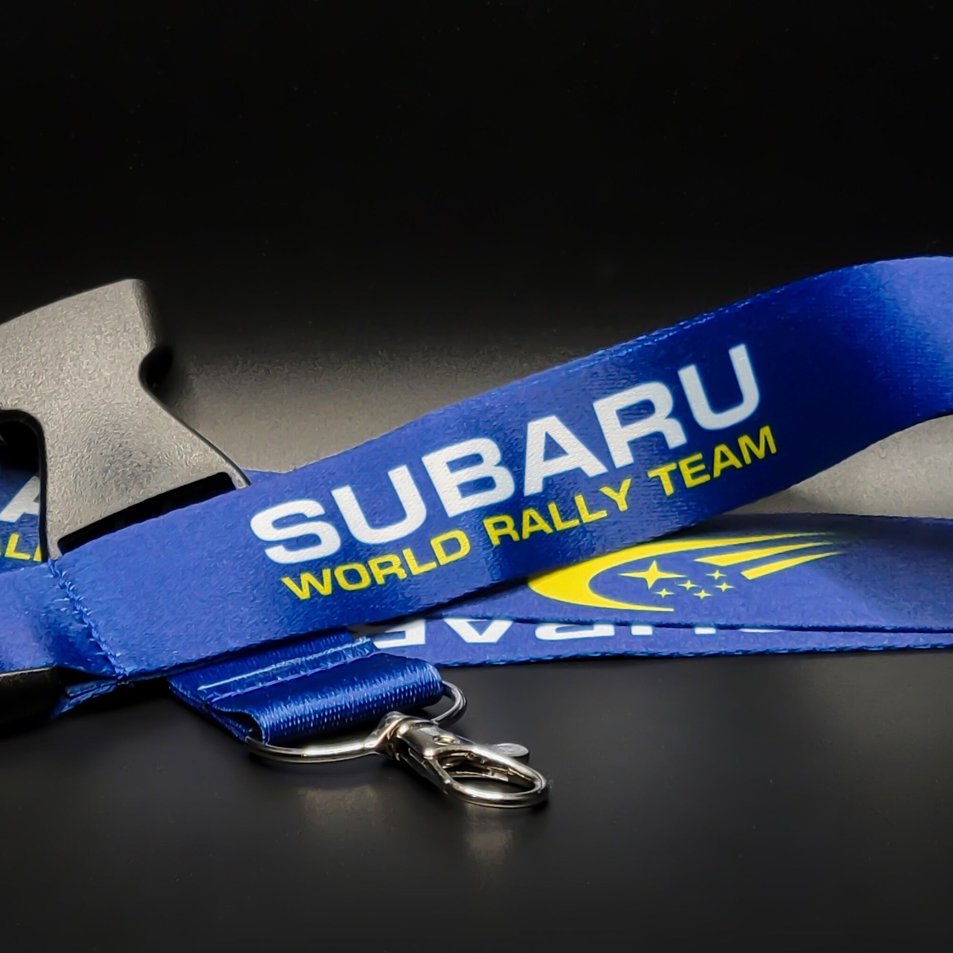 Blue Subaru lanyard with white Subaru and yellow World Rally Team text plus the yellow Subaru stars logo, coiled with a grey breakaway clip and silver clasp on black.