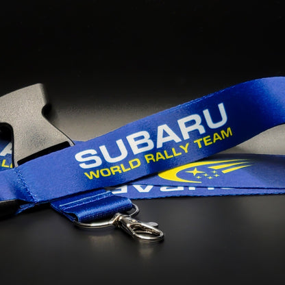 Blue Subaru lanyard with white Subaru and yellow World Rally Team text plus the yellow Subaru stars logo, coiled with a grey breakaway clip and silver clasp on black.