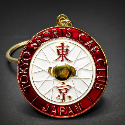 Double-sided round metal Tokyo Sports Car Club badge keychain, red ring with gold TOKYO SPORTS CAR CLUB and JAPAN lettering, white pinwheel center with gold kanji and a raised gold sphere, gold hanging loop.
