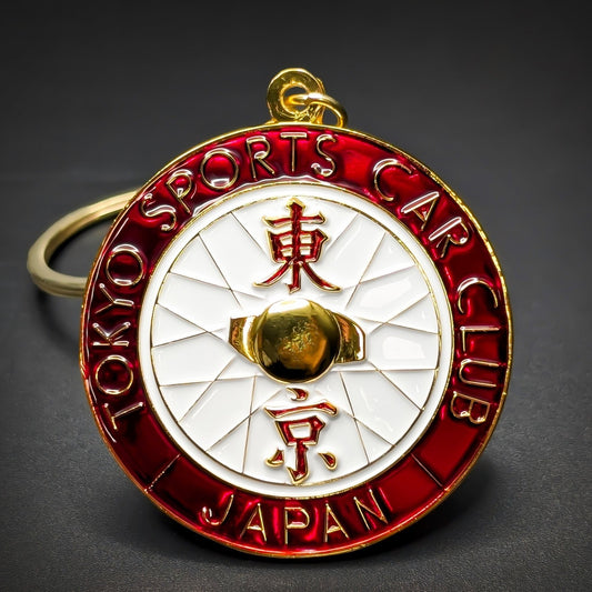 Double-sided round metal Tokyo Sports Car Club badge keychain, red ring with gold TOKYO SPORTS CAR CLUB and JAPAN lettering, white pinwheel center with gold kanji and a raised gold sphere, gold hanging loop.