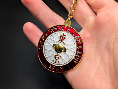 Hand holding a red and white Tokyo Sports Car Club enamel badge keychain to show size and metallic gold finish.