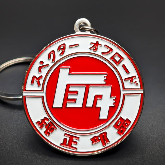 Double-sided metal Toyota JDM badge keychain, a red and white enamel medallion with Japanese lettering arched around the rim and a white emblem on the center disc, split ring, dark surface.