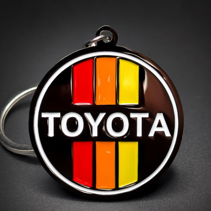 Double-sided metal Toyota badge keychain, black enamel medallion with a white circle outline, red, orange, and yellow stripes top and bottom, and white “TOYOTA” lettering at center, silver split ring.