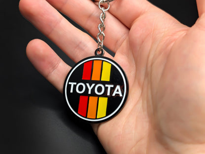 Hand holding a circular Toyota retro stripes enamel badge keychain to demonstrate the premium metallic weight and vibrant color finish.