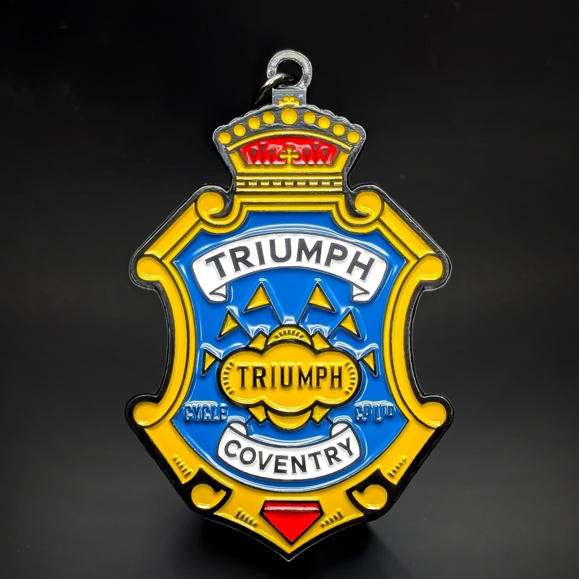 Double-sided metal Triumph badge keychain shaped like a gold crest, with a crown, a blue center panel, white ‘TRIUMPH’ and ‘COVENTRY’ banners, and a gold ‘TRIUMPH’ medallion, on a dark background.
