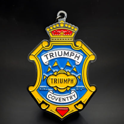 Double-sided metal Triumph badge keychain shaped like a gold crest, with a crown, a blue center panel, white ‘TRIUMPH’ and ‘COVENTRY’ banners, and a gold ‘TRIUMPH’ medallion, on a dark background.