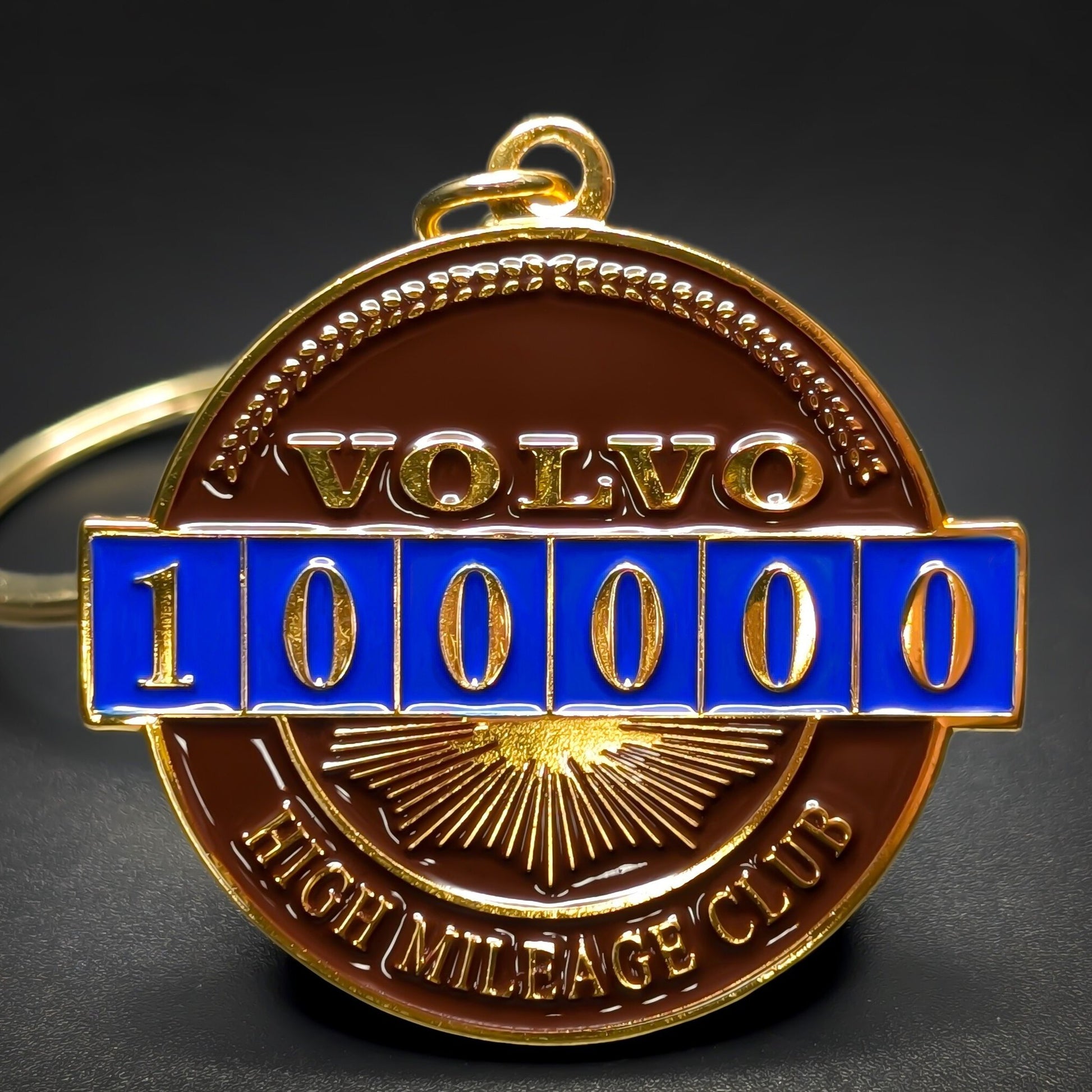 Double-sided metal Volvo badge keychain in gold-tone metal with VOLVO lettering above a blue enamel odometer reading 100000, HIGH MILEAGE CLUB text below, gold split ring, on a dark surface.