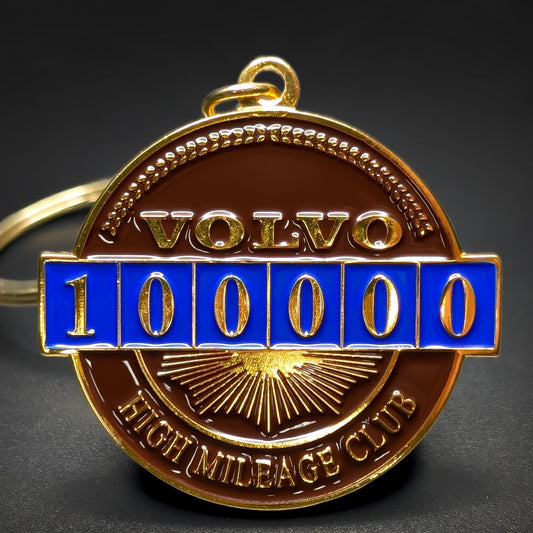 Double-sided metal Volvo badge keychain in gold-tone metal with VOLVO lettering above a blue enamel odometer reading 100000, HIGH MILEAGE CLUB text below, gold split ring, on a dark surface.
