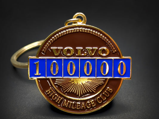 Close-up of a circular gold-tone metal badge keychain for the Volvo 100,000 High Mileage Club with brown and blue enamel.