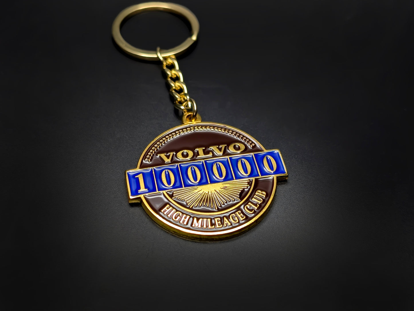 A Volvo 100k mileage badge keychain featuring a gold chain and ring, laid flat on a dark, professional surface.