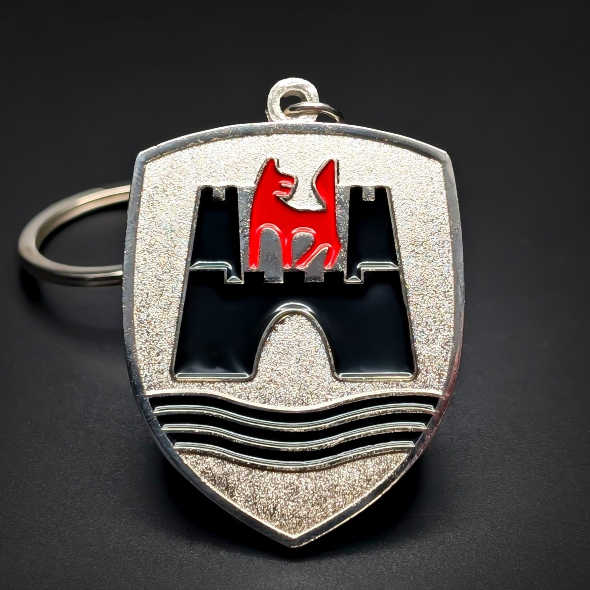 Double-sided shield-shaped metal Volkswagen Wolfsburg badge keychain, brushed silver face with a black enamel castle and a red wolf emblem above black wave lines, chrome border, chain and split ring, dark background.