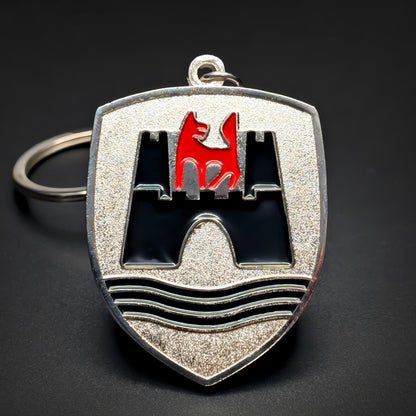 Double-sided shield-shaped metal Volkswagen Wolfsburg badge keychain, brushed silver face with a black enamel castle and a red wolf emblem above black wave lines, chrome border, chain and split ring, dark background.